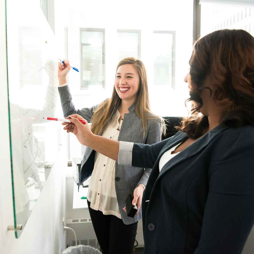 Two professional women collaborate enthusiastically at a whiteboard in a bright office. Both are smiling and engaged, reflecting a positive and collaborative work environment that promotes employee wellbeing through teamwork and open communication.