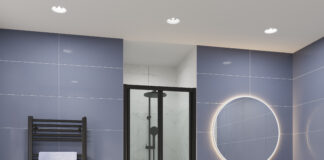 Modern bathroom with a compact black-framed glass shower enclosure in the center, surrounded by glossy blue wall tiles. On the left is a black towel radiator with a light blue towel, and on the right is a white floating vanity with black handles, a black faucet, and a round LED-lit mirror above it. The floor is tiled in light beige.